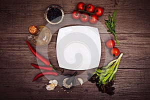 Fresh tomatoes, chili pepper and other spices and herbs around modern white square plate in the center of wooden table and cloth