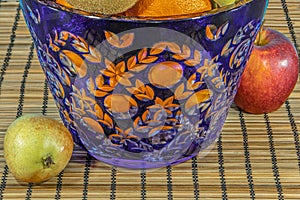 Fresh tropical and subtropical fruits in a large blue crystal vase. Wicker bamboo stand on the dining table. Close up macro