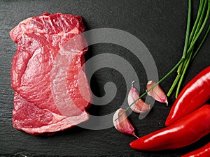 Fresh uncooked raw sirloin steak, red chilli peppers garlic cloves and herbs on a dark stone board. Still life