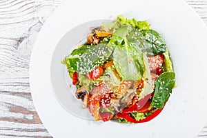 Fresh vegetable salad with lettuce, spinach, grilled mushrooms, tomatoes, sweet peppers and sesame seeds on plate on light wooden