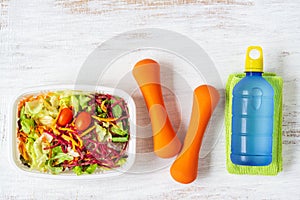 Fresh vegetable salad in lunch box with orange dumbbells excercise equipment and energy water drink on white rusty wood background