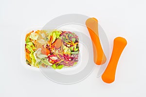 Fresh vegetable salad in lunch box with orange dumbbells excercise equipment on white. Active healthy lifestyles, good health