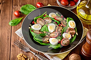 Fresh vegetable salad with spinach, cherry tomatoes, quail eggs, pomegranate seeds and walnuts in black plate on wooden table.