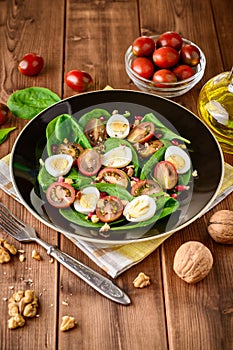 Fresh vegetable salad with spinach, cherry tomatoes, quail eggs, pomegranate seeds and walnuts in black plate on wooden table.