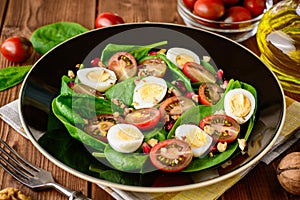 Fresh vegetable salad with spinach, cherry tomatoes, quail eggs, pomegranate seeds and walnuts in black plate on wooden table.