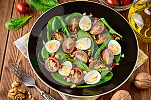 Fresh vegetable salad with spinach, cherry tomatoes, quail eggs, pomegranate seeds and walnuts in black plate on wooden table.