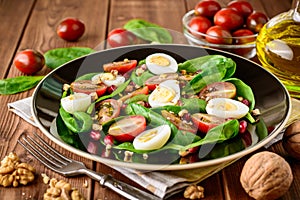 Fresh vegetable salad with spinach, cherry tomatoes, quail eggs, pomegranate seeds and walnuts in black plate on wooden table.