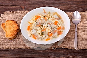 Fresh vegetable soup with pieces of chicken breast and noodles in a white plate served with slices of toast, angle view