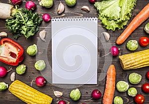 Fresh vegetables and herbs laid out around a notebook for recipes on wooden rustic background top view close up border, place for