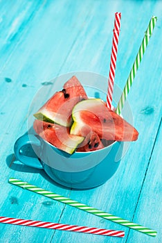 Fresh watermelon slices in a cup with coloured straws, blue wooden background