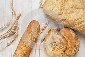 Fresh white wheat bread, round classic, ciabatta, french baguette, wheat ears and grains on white wooden background top view.