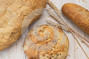 Fresh white wheat bread, round classic, ciabatta, french baguette, wheat ears and grains on white wooden background top view.