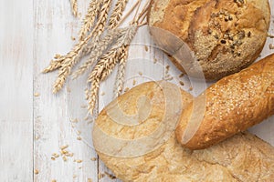 Fresh white wheat bread, round classic, ciabatta, french baguette, wheat ears and grains on white wooden background top view.