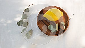 Fresh whole lemon fruit on wooden plate and dried eucalyptus twigs on linen tablecloth in sunlight