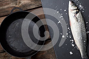 Fresh whole sea bass fish on a black board, next to it is an empty round black frying pan