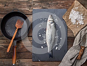 fresh whole sea bass fish on a black graphite board, next to it is an empty round black frying pan