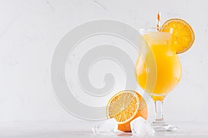 Fresh yellow oranges cocktail in elegance wineglass with ice cubes, straw and half oranges on soft white wood background.