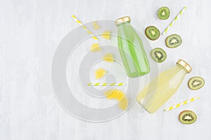 Fresh yellow pineapple and green kiwi juices in glass bottles with sliced fruit, straws on white wooden board, top view.