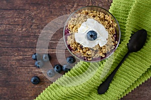 Freshly baked blueberry crunch in a glass bowl on a rustic wood table, whipped cream, green towel, black spoon