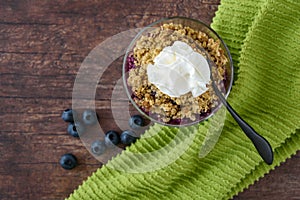 Freshly baked blueberry crunch in a glass bowl on a rustic wood table, whipped cream, green towel, black spoon