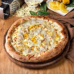 Freshly baked crust Hawaiian pizza ham and pineapple on a wooden board surrounded by ingredients in individual.