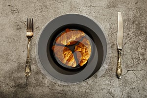 Freshly baked French croissants on a black plate with a fork and knife on a gray concrete background. View from above.