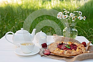 Freshly baked homemade galette or open strawberry pie, a cup of herbal tea, a teapot and a vase with a bouquet of daisy flowers,