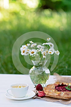 Freshly baked homemade galette or open strawberry pie, a cup of herbal tea and a vase with a bouquet of daisy flowers. Soft