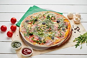 Freshly baked pizza with ham, rukkola, sauce pesto and olives served on wooden background with tomatoes, sauces and herbs. Food