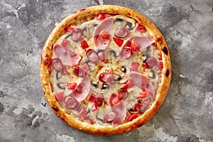 Freshly baked pizza with hunting sausages, ham, mushrooms and tomatoes on gray stone surface