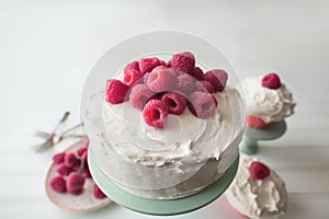 Freshly baked raspberry cake with white frosting and fresh raspberries, two cupcakes in the background.