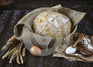 Freshly baked rye bread with flax seeds in a burlap basket, wooden spoons with flour, wheat ears, egg.