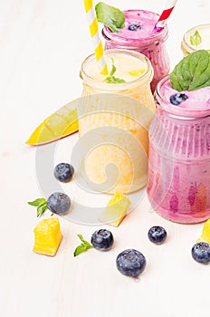 Freshly blended yellow and violet fruit smoothie in glass jars with straw, mint leaves, mango slices, blueberry, close up. Soft w