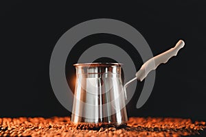 Freshly brewed coffee in cezve on a dark golden background next to scattered roasted coffee beans