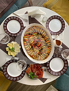 Freshly cooked ratatouille stands in the center of a round table set with jamon, cheese and sauce.