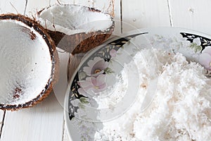 Freshly grated coconut on plate and coconut shell on white wooden table