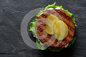 Freshly grilled plant based burger patty on bun with lettuce, slices of gherkin and sauce isolated on black slate. Top view. Copy