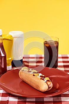 Freshly made hot dog with mustard and ketchup on a plate with coke glass on red gingham table cloth over yellow