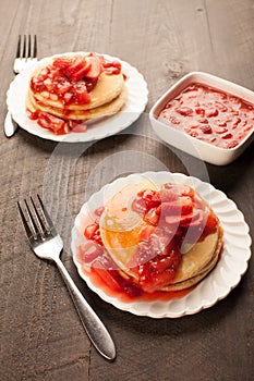 Freshly made white flour pancakes stacked on white plates topped with red strawberry syrup vertical shot