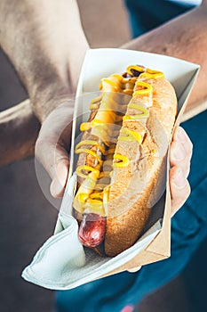 Freshly prepared cheesy hotdog in a paper box in a hand. Food delivery or lyfestyle concept
