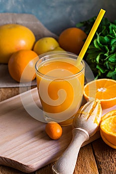 Freshly pressed citrus fruit juice from oranges lemons limes cumquat, glass with straw, reamer, mint on table by window, rustic