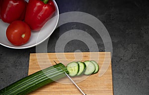 Freshly sliced cucumbers with knife on wooden cutting board with a plate of paprika and tomato