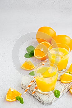 Freshly squeezed orange juice with ice in a glass with a straw on a wooden board on a light background with fresh oranges.