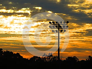 Friday night lights during the time of COVID: Playing field floodlights against clouds and trees at sunset