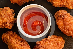 Fried breaded chicken strips with red sauce on a black table. View from above