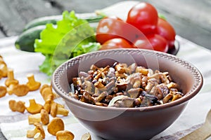 Fried chanterelles with onion in rustic bowl and plate with fresh vegetables for salad on background