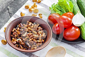 Fried chanterelles with onion in rustic bowl and plate with fresh vegetables for salad on background