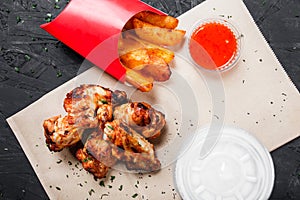 Fried chicken wings with baked potatoes and bbq sauce on craft paper on black background