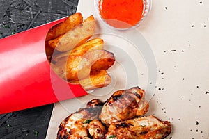 Fried chicken wings with baked potatoes and bbq sauce on craft paper on black background.