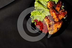 fried chicken wings with salad and ketchup in a black plate and fork on black background.